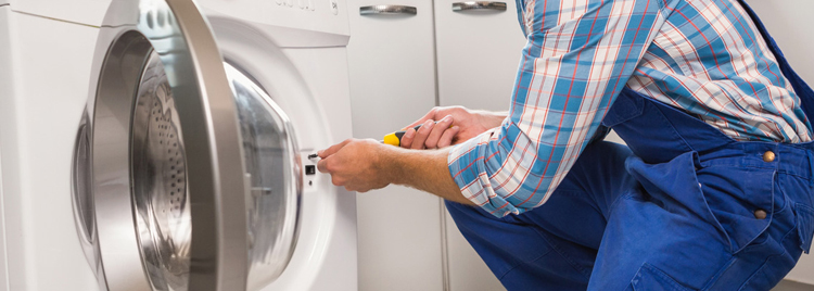 An appliance repair technician in blue overalls fixing a washer/dryer