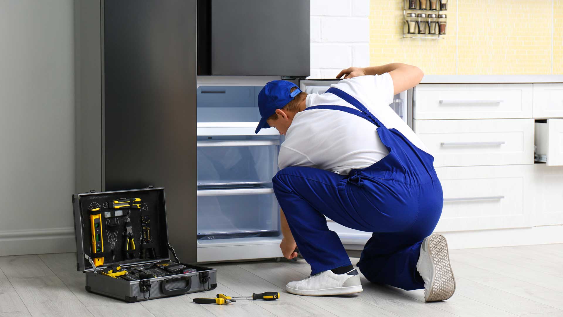 A man in blue overalls repairing a refrigerator